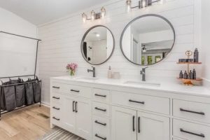 Close-up of white shaker double vanity with black hardware, twin round mirrors, and brass sconces on a shiplap wall.